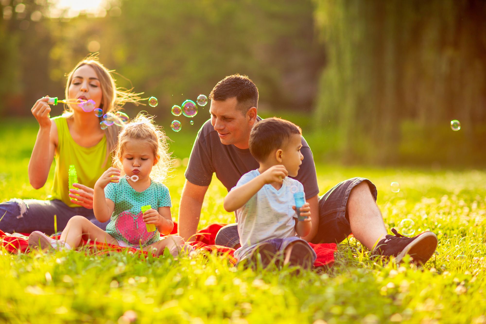 Parents and their two young children blow bubbles together on the grass in late-afternoon light