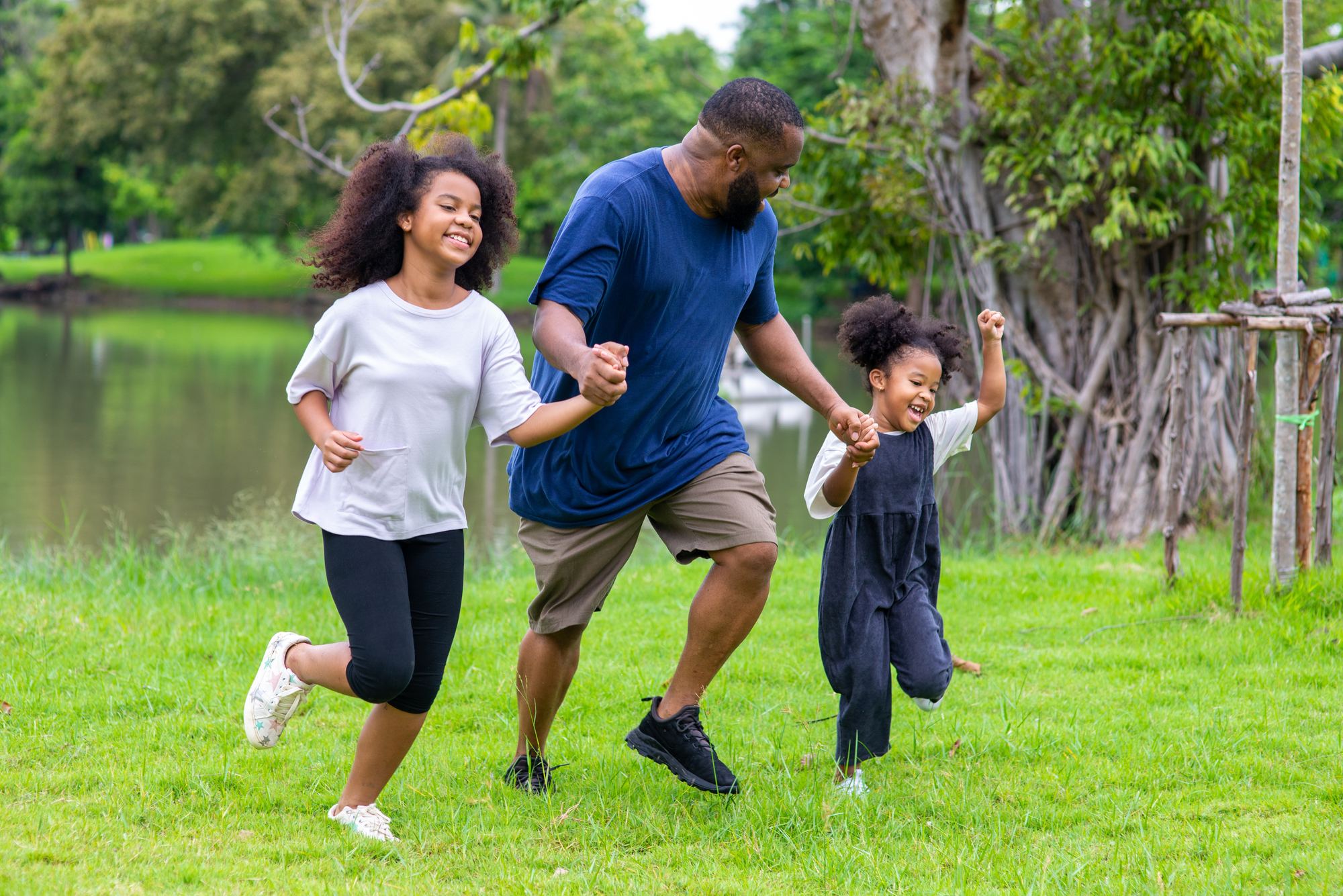 A father runs hand-in-hand through a park with his two laughing daughters